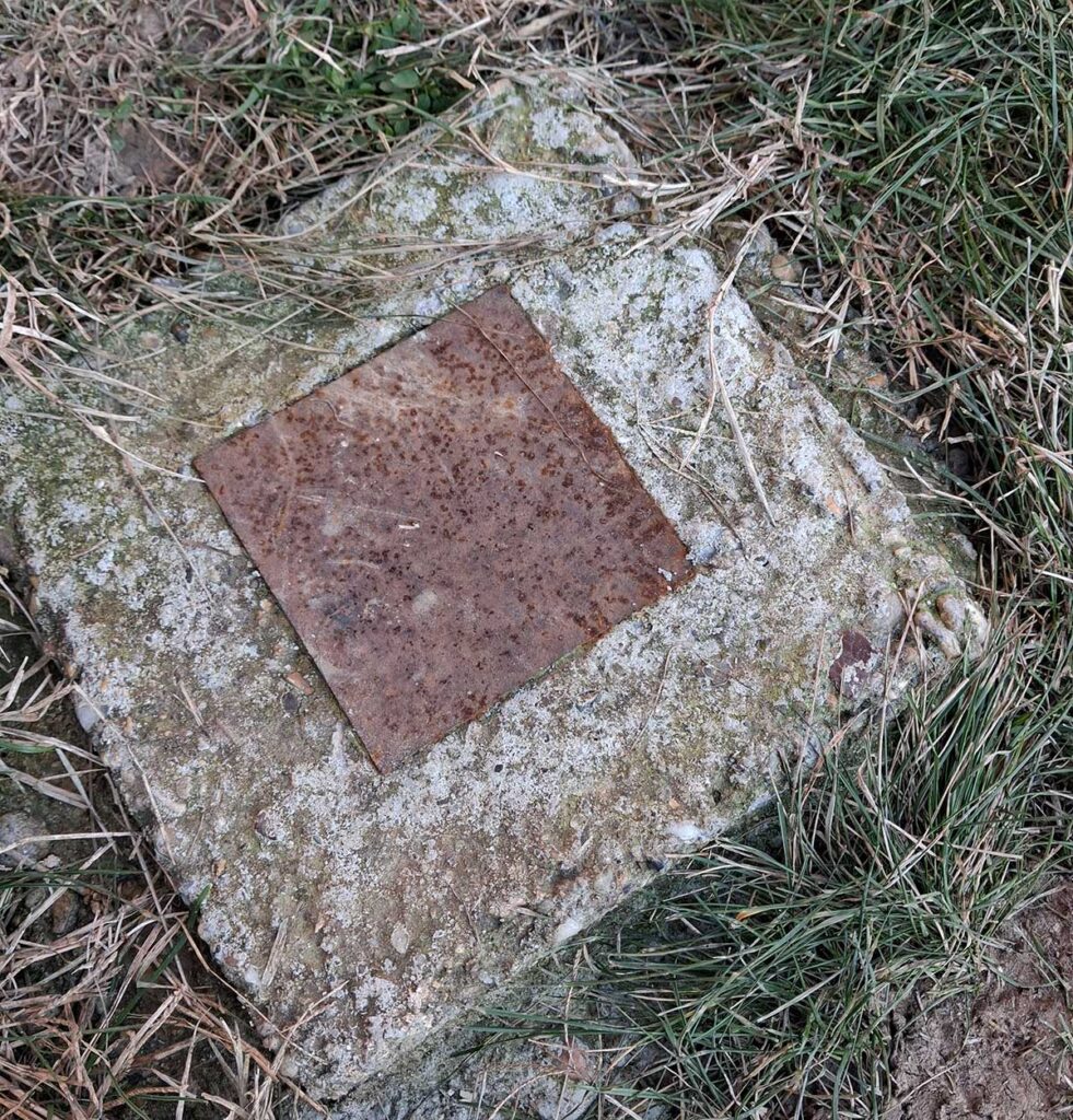 Real photo of a metal anchor plate embedded in a fresh concrete footing, with welded metal keys locked into the concrete for strong attachment on a sloped deck foundation.