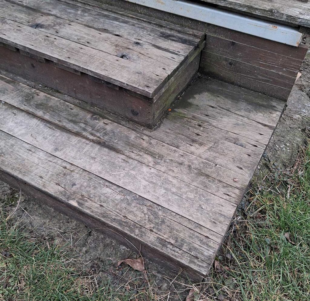 Close-up of a two-step metal staircase for a deck on slope: Welded rectangular tubing stringers clad with matching wood decking boards for safe access down the elevation change.